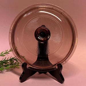 May include: A vintage pink glass lid with a dark brown knob, displayed on a black stand. The lid features a radial design emanating from the center knob. The glass has a slight tint, and the lid is round.
