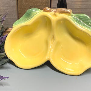 May include: A ceramic serving dish in the shape of two pears, painted in a bright yellow color. The dish has green leaves and a brown stem. The pear-shaped bowls are ideal for serving snacks or dips.