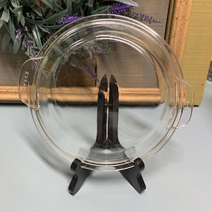 Könnte beinhalten: Eine transparente Pyrex-Tarteform mit Griffen, auf einem schwarzen Präsentationsständer. Die runde Form scheint leer zu sein. Im Hintergrund ein dekorativer Kranz mit grünen Blättern und lila Blüten. Durchmesser ca. 23 cm.