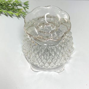 Könnte beinhalten: Ein klarer, antiker Kerzenhalter aus Glas mit einem Rautenmuster auf dem Sockel und einem gewellten Rand. Der Kerzenhalter steht auf dem Kopf und zeigt sein kompliziertes Design und seine filigranen Details. Ein Zweig Grünzeugs ist im Hintergrund.