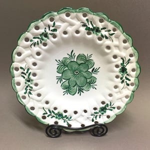 May include: White ceramic plate with a green floral design in the centre and around the rim. The plate has a woven pattern with small holes and a scalloped edge. The plate is displayed on a black metal stand.
