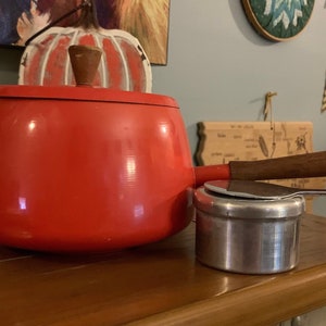 May include: A bright orange fondue pot with a wooden handle and a silver metal base. The pot is on a wooden surface.