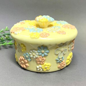 May include: A yellow ceramic trinket box with a lid, adorned with pastel-colored floral embellishments. The box features a round shape with a matching floral knob on the lid. The flowers are in shades of yellow, pink, and blue.