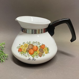 May include: White enamel tea kettle with a black handle and a silver band. The kettle features a colorful floral and fruit design with the words "Le Thé" in black script. The kettle has a spout for pouring.