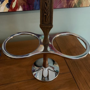 Könnte beinhalten: Ein doppelter Flaschenhalter aus Chrom und Holz mit einem geschnitzten Blumenmuster auf dem Holzgriff. Der Halter hat zwei runde Öffnungen für Flaschen und einen runden Sockel.