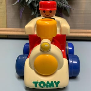 May include: A red, yellow, and blue toy train with a smiling figure in a red hat driving it. The train has the word "TOMY" printed on the front.