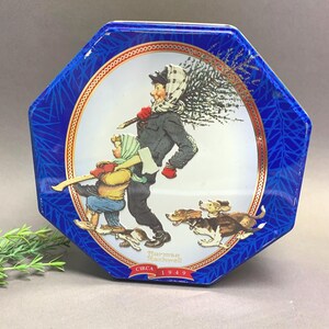 Könnte beinhalten: Achteckige blaue Dose mit dekorativer Illustration. Die Illustration zeigt eine Person, die einen Weihnachtsbaum trägt, gefolgt von einem Kind und Hunden. Die Dose hat einen weißen Innenraum mit goldenem Rand und dem Text "Norman Rockwell Circa 1949".