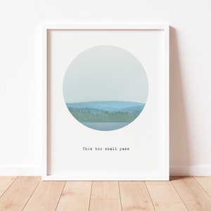 Peut inclure: Une impression encadrée avec un fond blanc et un cercle au centre. Le cercle contient une photo d'un lac et d'arbres avec un ciel bleu. Le texte "This too shall pass" est imprimé sous le cercle.