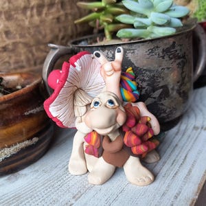 Puede incluir: Una caprichosa figura de arcilla polimérica de una criatura parecida a una tortuga con un sombrero de hongo y una mano haciendo el signo de la paz. La figura tiene un cuerpo beige, un chaleco marrón y una bufanda colorida. La seta es roja con lunares blancos.