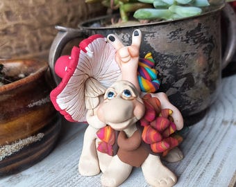 Puede incluir: Una caprichosa figura de arcilla polimérica de una criatura parecida a una tortuga con un sombrero de hongo y una mano haciendo el signo de la paz. La figura tiene un cuerpo beige, un chaleco marrón y una bufanda colorida. La seta es roja con lunares blancos.