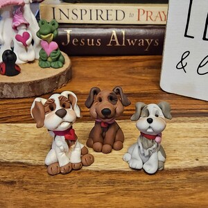Puede incluir: Tres figuras de perros hechas a mano. Una es blanca y marrón, otra es marrón y la tercera es gris y blanca. Cada perro tiene un lazo rojo. Las figuras están sobre una superficie de madera.