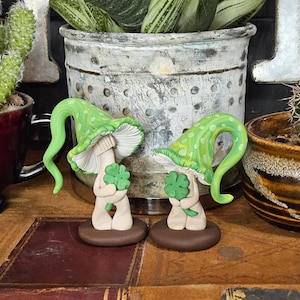 May include: Two whimsical mushroom figurines with green polka-dotted caps and white undersides, each holding a green four-leaf clover. The mushroom stems are beige, and the bases are brown. The figurines are set on a wooden surface.