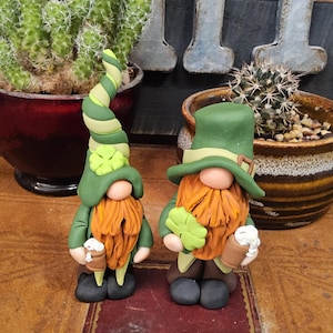Puede incluir: Dos gnomos del Día de San Patricio. Uno tiene un sombrero verde y amarillo en espiral, el otro un sombrero verde tradicional. Ambos tienen barbas naranjas, tréboles verdes y sostienen jarras de cerveza. Están hechos de arcilla.