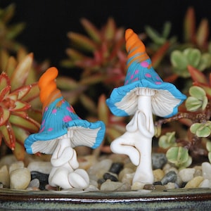 Puede incluir: Dos figuritas de champiñones de cerámica con sombreros azules y rosas y cuerpos blancos en una postura de yoga. Los champiñones están sentados en una cama de pequeñas piedras.