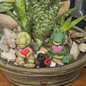 Puede incluir: Una maceta de cerámica con una planta de aloe vera, pequeñas rocas y figuras decorativas. Las figuras incluyen una tortuga y una rana sosteniendo corazones, y una mariquita. La maceta es de color verde apagado.