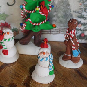 May include: Three handmade Christmas decorations: a gingerbread person with a candy cane scarf, a snowman with a blue and white scarf, and a snowman with a green and white scarf. Both snowmen have red robins on their heads.
