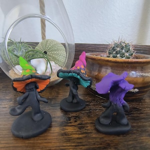 May include: Three whimsical clay mushroom figurines with colorful hats. One has a green and orange hat, another a pink and teal hat, and the third a purple hat. Each mushroom has a black stem and base, and they are displayed on a wooden surface.