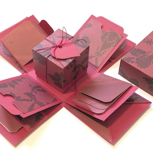 May include: A handmade explosion box in shades of red and brown. The box is open, revealing multiple layers with pockets and cards. The exterior features a damask pattern, and the central cube is tied with string and a heart-shaped tag.