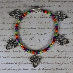 Può includere: Un braccialetto multi-colorato con ciondoli a forma di drago argentati. Le perline sono arancioni, rosse, verdi, gialle e viola.