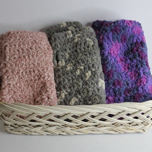 May include: Three crocheted blankets in a wicker basket. The blankets are pink, gray with white flecks, and purple with pink flecks.