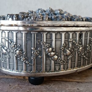 May include: A silver oval-shaped trinket box with a detailed floral design and a keyhole on the side. The box has a raised lid with a floral design and sits on four small black feet.