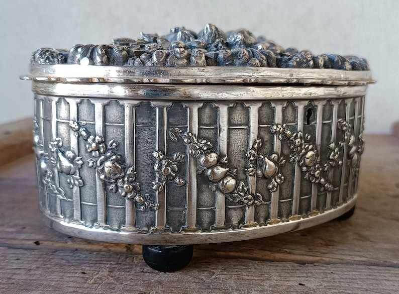 May include: A silver oval-shaped trinket box with a detailed floral design and a keyhole on the side. The box has a raised lid with a floral design and sits on four small black feet.