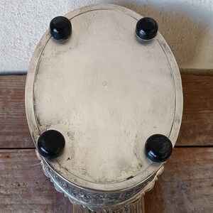 May include: Silver oval trinket box with a decorative, scalloped edge and four black feet. The box is closed and the top is smooth and reflective.