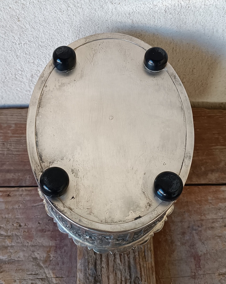 May include: Silver oval trinket box with a decorative, scalloped edge and four black feet. The box is closed and the top is smooth and reflective.