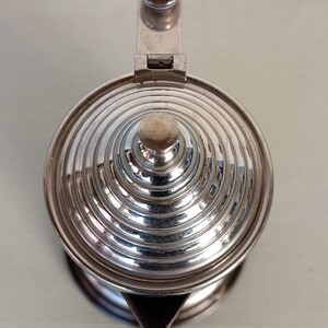 Peut inclure: Une th&eacute;i&egrave;re argent&eacute;e avec un couvercle conique &agrave; rainures et une petite poign&eacute;e ronde. Le bec verseur est triangulaire. La th&eacute;i&egrave;re est en m&eacute;tal brillant, refl&eacute;tant la lumi&egrave;re. Le couvercle a un petit bouton rond. La th&eacute;i&egrave;re est probablement utilis&eacute;e pour servir le th&eacute;.