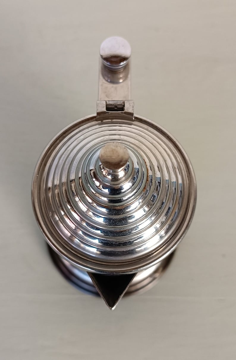 Peut inclure: Une th&eacute;i&egrave;re argent&eacute;e avec un couvercle conique &agrave; rainures et une petite poign&eacute;e ronde. Le bec verseur est triangulaire. La th&eacute;i&egrave;re est en m&eacute;tal brillant, refl&eacute;tant la lumi&egrave;re. Le couvercle a un petit bouton rond. La th&eacute;i&egrave;re est probablement utilis&eacute;e pour servir le th&eacute;.