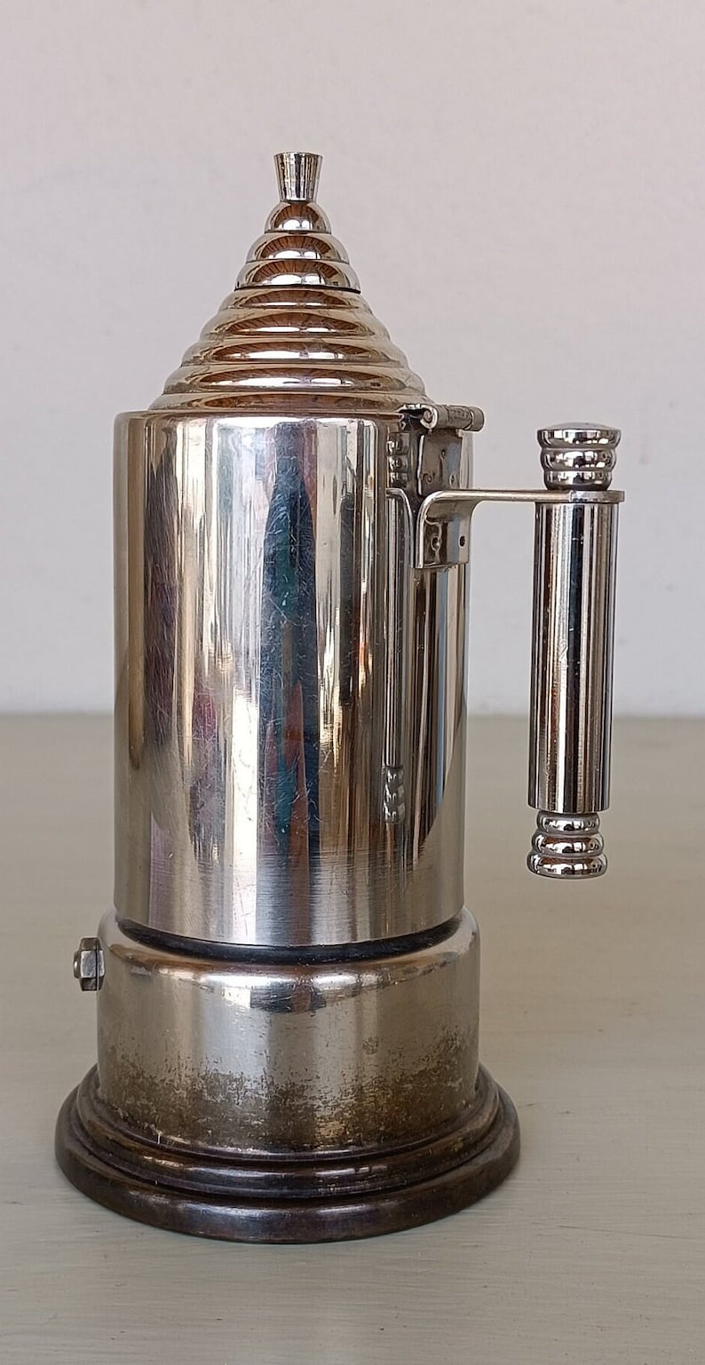 Peut inclure: Une cafeti&egrave;re vintage argent&eacute;e avec un corps cylindrique et un dessus conique. La poign&eacute;e et le dessus sont orn&eacute;s de motifs c&ocirc;tel&eacute;s. La base est marron fonc&eacute;. La cafeti&egrave;re est probablement en m&eacute;tal et pr&eacute;sente un design classique.