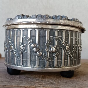 May include: A silver trinket box with an ornate design featuring flowers and fruit. The box has a hinged lid and rests on three small feet.