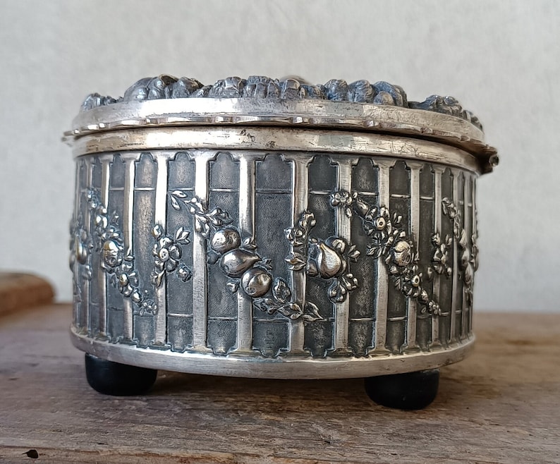 May include: A silver trinket box with an ornate design featuring flowers and fruit. The box has a hinged lid and rests on three small feet.