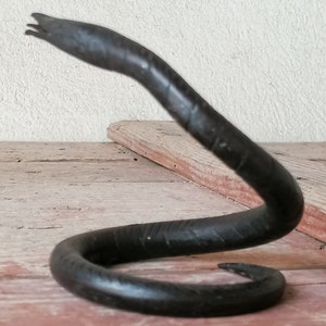 K&ouml;nnte beinhalten: Eine schwarze Schlangenskulptur aus Metall mit einem gewundenen K&ouml;rper und einem erhobenen Kopf. Die Skulptur steht auf einer Holzoberfl&auml;che.