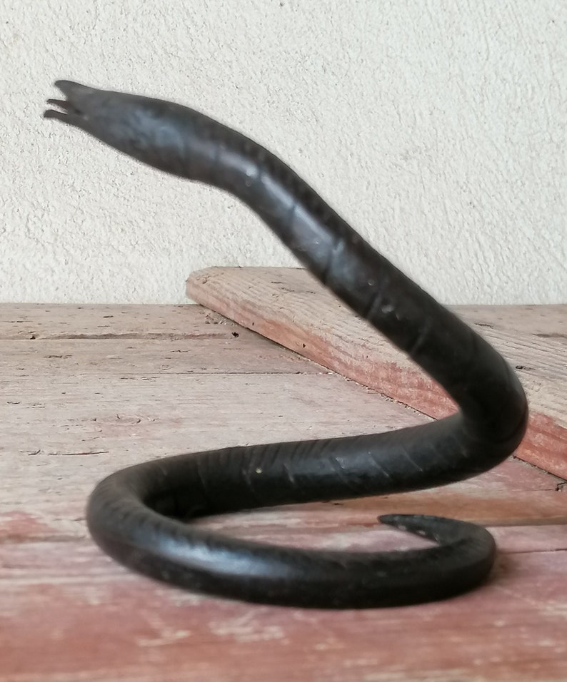 K&ouml;nnte beinhalten: Eine schwarze Schlangenskulptur aus Metall mit einem gewundenen K&ouml;rper und einem erhobenen Kopf. Die Skulptur steht auf einer Holzoberfl&auml;che.