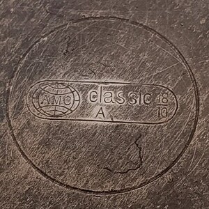Peut inclure: Gros plan d'une surface m&eacute;tallique avec le texte en relief "AMC classic 18/10" dans un cadre circulaire. Le design comprend un logo de globe et la lettre "A". La surface pr&eacute;sente des rayures visibles.