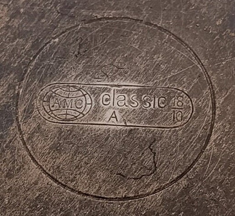 Peut inclure: Gros plan d'une surface m&eacute;tallique avec le texte en relief "AMC classic 18/10" dans un cadre circulaire. Le design comprend un logo de globe et la lettre "A". La surface pr&eacute;sente des rayures visibles.