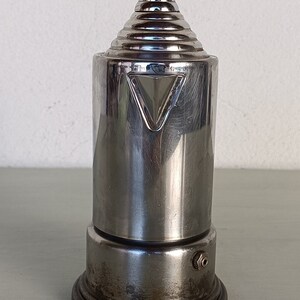 Peut inclure: Une cafeti&egrave;re vintage en m&eacute;tal argent&eacute; avec un dessus conique et un corps cylindrique. Le design pr&eacute;sente une s&eacute;rie d'anneaux concentriques sur le dessus et un accent triangulaire sur le corps. La base est de forme circulaire et sombre.