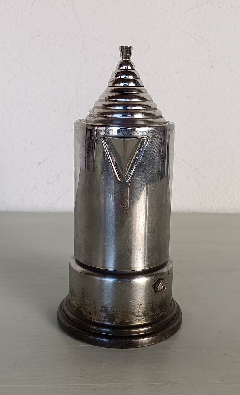 Peut inclure: Une cafeti&egrave;re vintage en m&eacute;tal argent&eacute; avec un dessus conique et un corps cylindrique. Le design pr&eacute;sente une s&eacute;rie d'anneaux concentriques sur le dessus et un accent triangulaire sur le corps. La base est de forme circulaire et sombre.