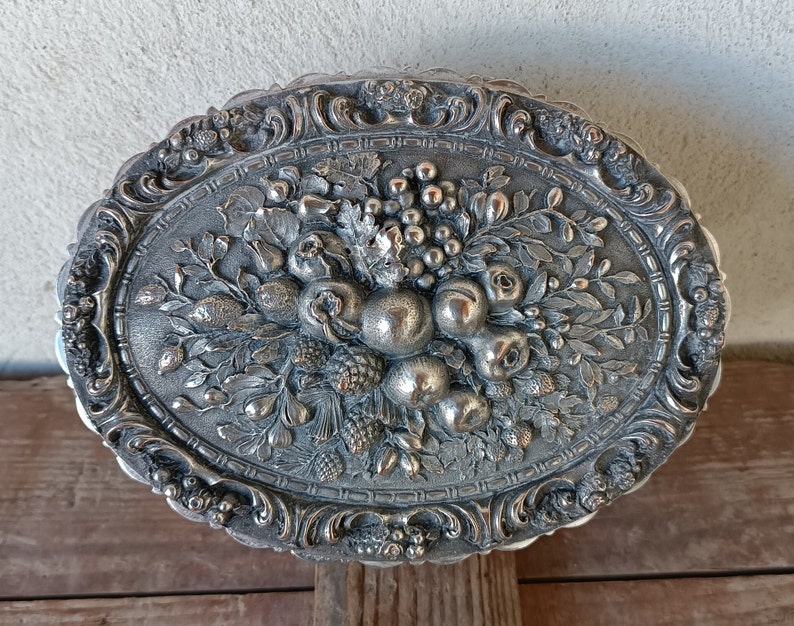 May include: Silver oval trinket box with an ornate design featuring a raised floral motif of fruit and leaves.