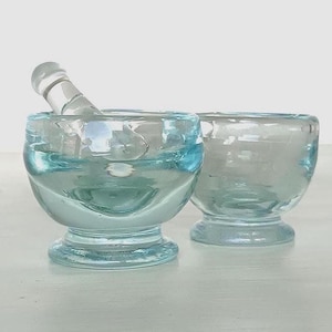 Op de afbeelding: Twee heldere, lichtblauwe glazen vijzel- en stamper sets. Eén set heeft de stamper in de vijzel. De vijzel en stamper zijn gemaakt van dik glas en hebben een afgeronde vorm. De andere vijzel is leeg.