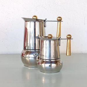 May include: Two silver and gold stovetop espresso makers. The larger maker has a gold handle and the smaller maker has a gold handle and a gold knob.