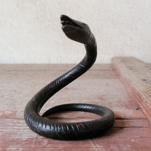 K&ouml;nnte beinhalten: Eine schwarze Schlangenskulptur aus Metall mit einem gewundenen K&ouml;rper und einem erhobenen Kopf. Die Schlange ist mit strukturierten Schuppen und einem leicht ge&ouml;ffneten Maul detailliert.
