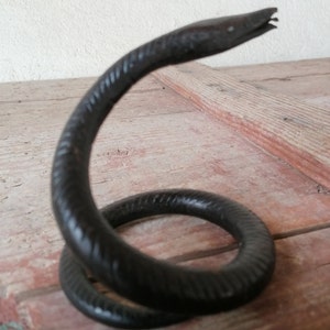 K&ouml;nnte beinhalten: Eine schwarze Schlangenskulptur aus Metall mit einem gewundenen K&ouml;rper und einem erhobenen Kopf. Die Schlange ist texturiert und hat ein realistisches Aussehen.