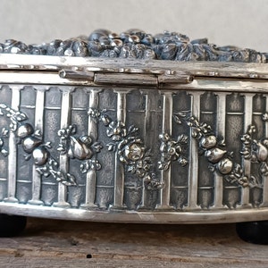 May include: Silver oval trinket box with an ornate floral design and a hinged lid. The box has a textured surface and sits on four small feet.