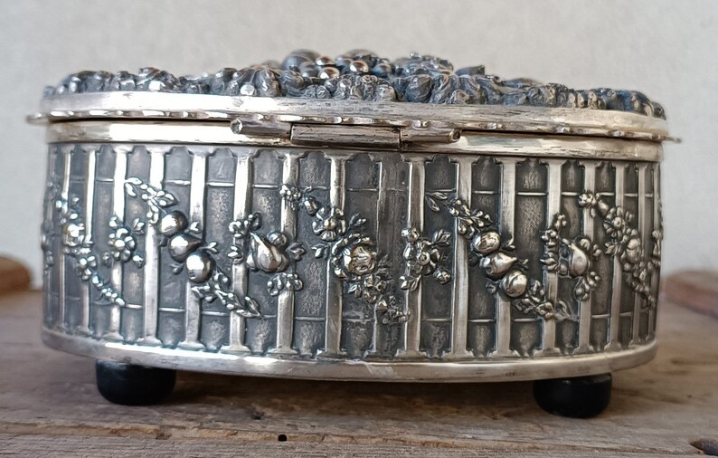 May include: Silver oval trinket box with an ornate floral design and a hinged lid. The box has a textured surface and sits on four small feet.