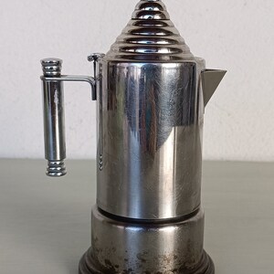 Peut inclure: Une cafeti&egrave;re italienne vintage en acier inoxydable. La cafeti&egrave;re a un corps cylindrique, un couvercle &agrave; gradins et une poign&eacute;e lat&eacute;rale. La base est sombre et la cafeti&egrave;re montre des signes d'usure. Le bec verseur est triangulaire.