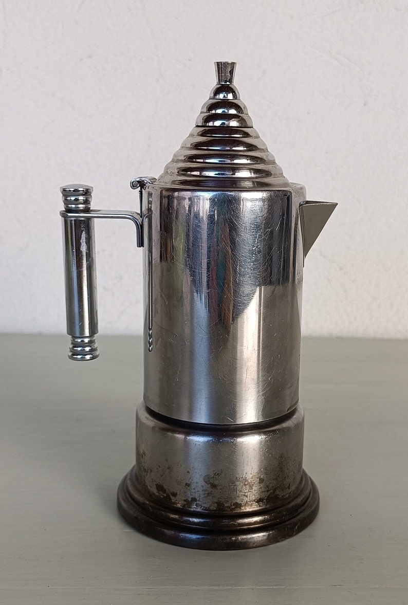Peut inclure: Une cafeti&egrave;re italienne vintage en acier inoxydable. La cafeti&egrave;re a un corps cylindrique, un couvercle &agrave; gradins et une poign&eacute;e lat&eacute;rale. La base est sombre et la cafeti&egrave;re montre des signes d'usure. Le bec verseur est triangulaire.