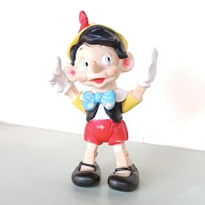 Könnte beinhalten: Eine Vintage-Pinocchio-Figur aus Plastik mit einem gelben Hut, schwarzen Haaren, einer blauen Fliege und roten Shorts. Die Figur hat eine lange Nase und hält die Arme in die Luft.