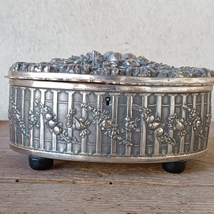 May include: A silver oval trinket box with an ornate floral design and a keyhole on the side. The box has three feet and is sitting on a wooden surface.